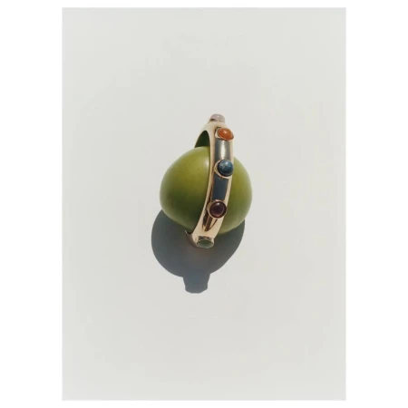 A gold ring with inset colored gemstones resting upright on a green olive against a clean white background.