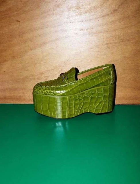 Platform shoe with alligator skin texture in bright green, set against a neutral wood panel and vibrant green surface.