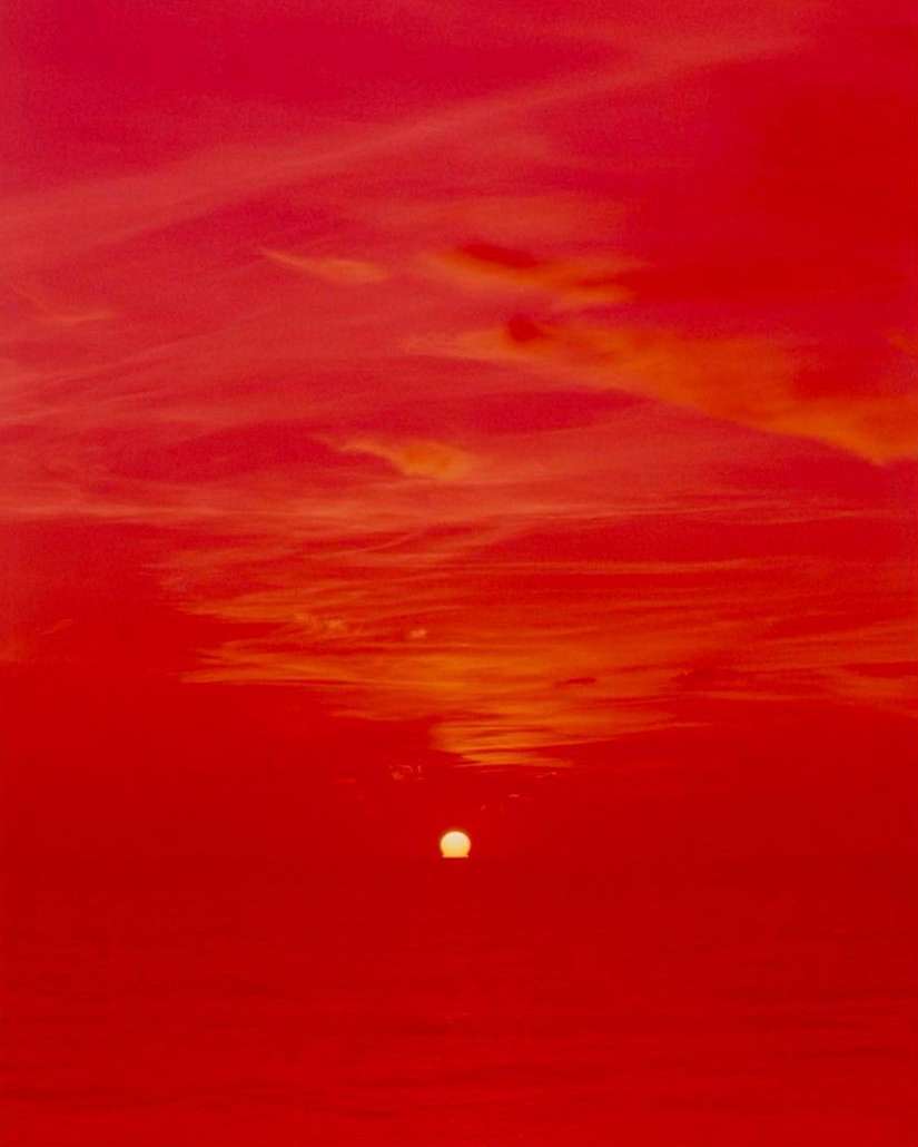 A vibrant red sky with clouds and a setting sun creates a striking landscape image, with the sun appearing as a bright, focal point.