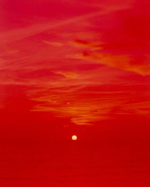 A vibrant red sky with clouds and a setting sun creates a striking landscape image, with the sun appearing as a bright, focal point.