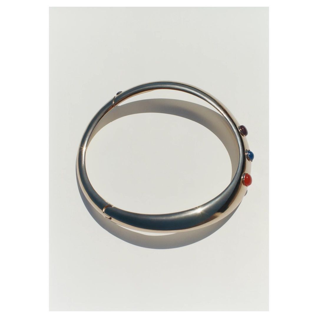 Gold bangle bracelet with red, purple and blue stones accents displayed on a white surface. Jewellery photography.