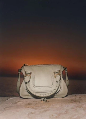 Light grey leather handbag sits on a stone surface against a gradient orange to dark grey background, showcasing fashion accessory.