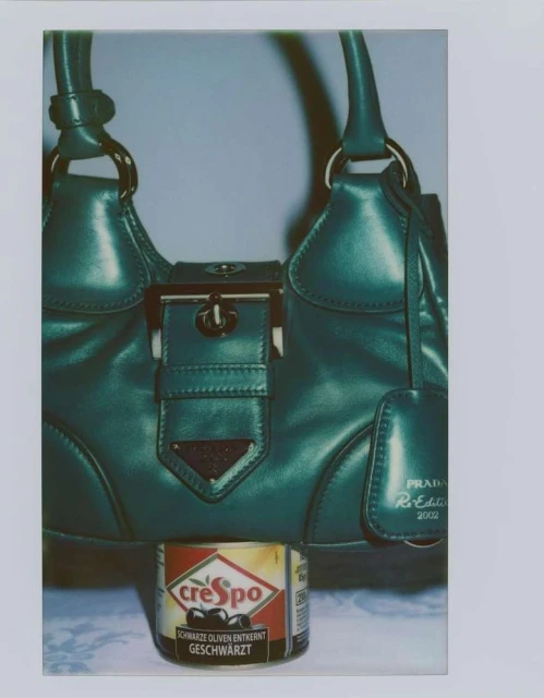 Green Prada handbag with silver hardware and a Crespo olive can in front, set against a soft grey background with patterned white cloth.