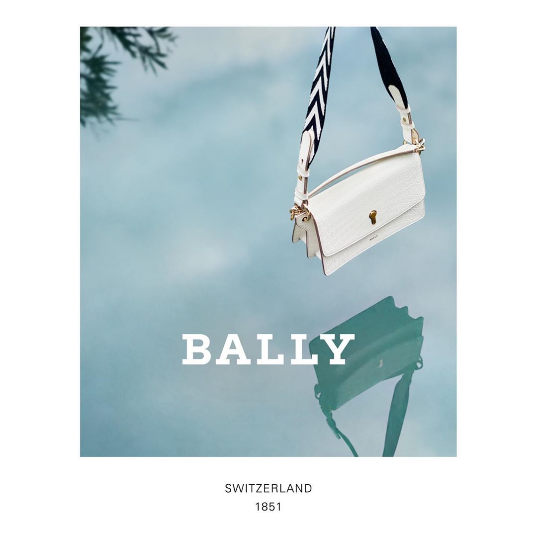 White leather Bally handbag with a black and white chevron strap, suspended against a gradient blue sky, with a mirrored reflection.