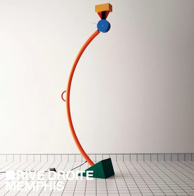 Memphis-style floor lamp with an orange curved stand, a blue disc, and a yellow cube sits on a tiled floor against a white wall.