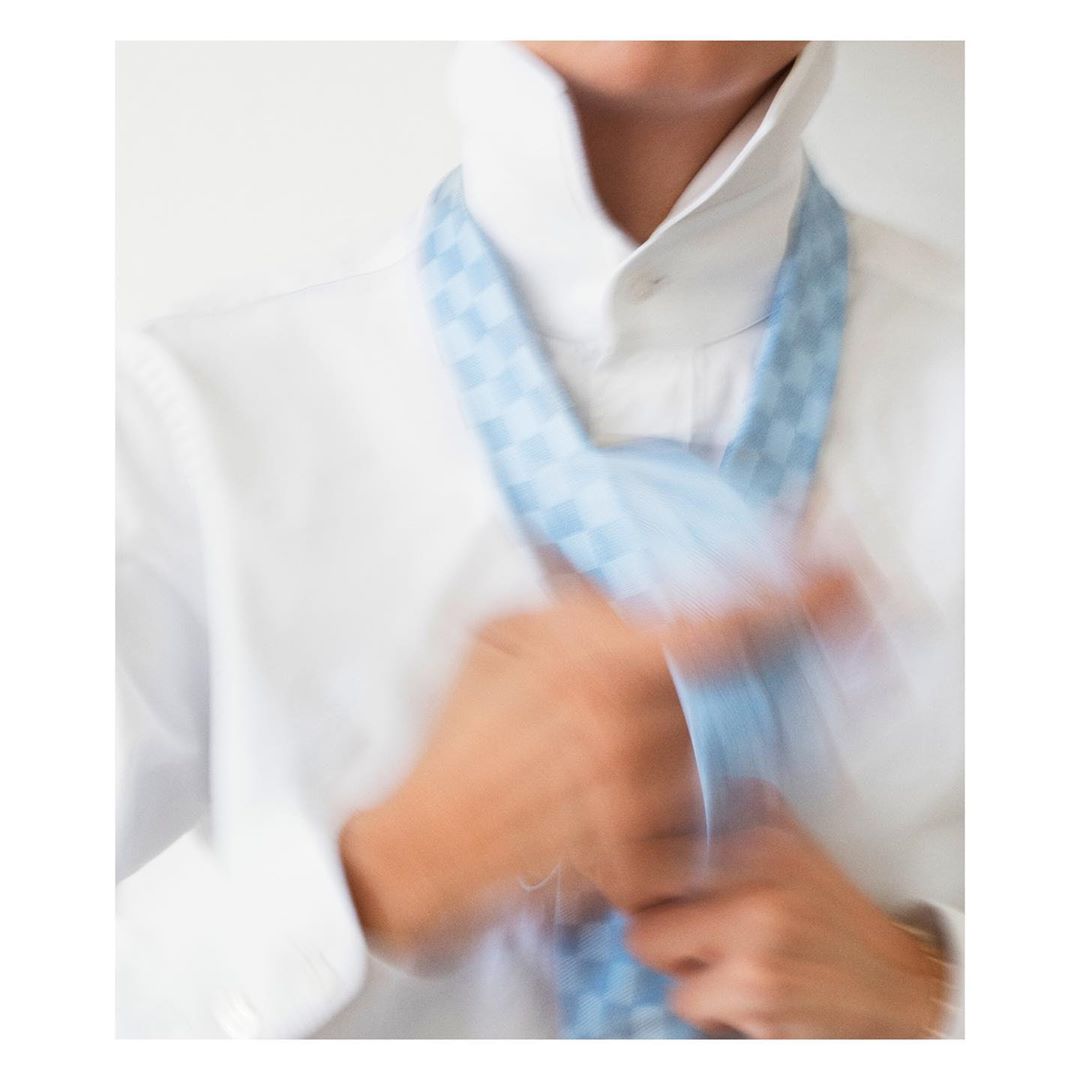 Man in a white shirt and blue checkered tie adjusting tie. The photo is slightly blurred, imparting a sense of motion. Indoor setting.