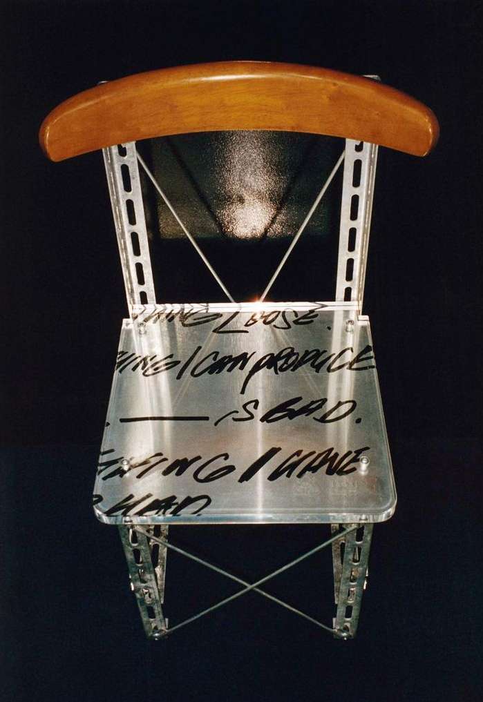 Unique art chair with metal frame, light wood back, and text across the clear seat, set against a stark black background.