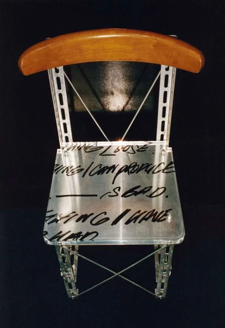 Unique art chair with metal frame, light wood back, and text across the clear seat, set against a stark black background.