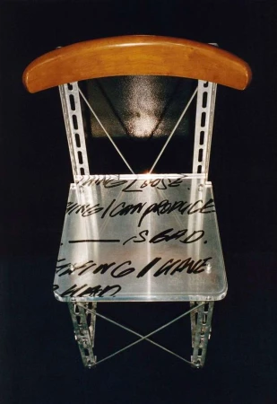 Unique art chair with metal frame, light wood back, and text across the clear seat, set against a stark black background.