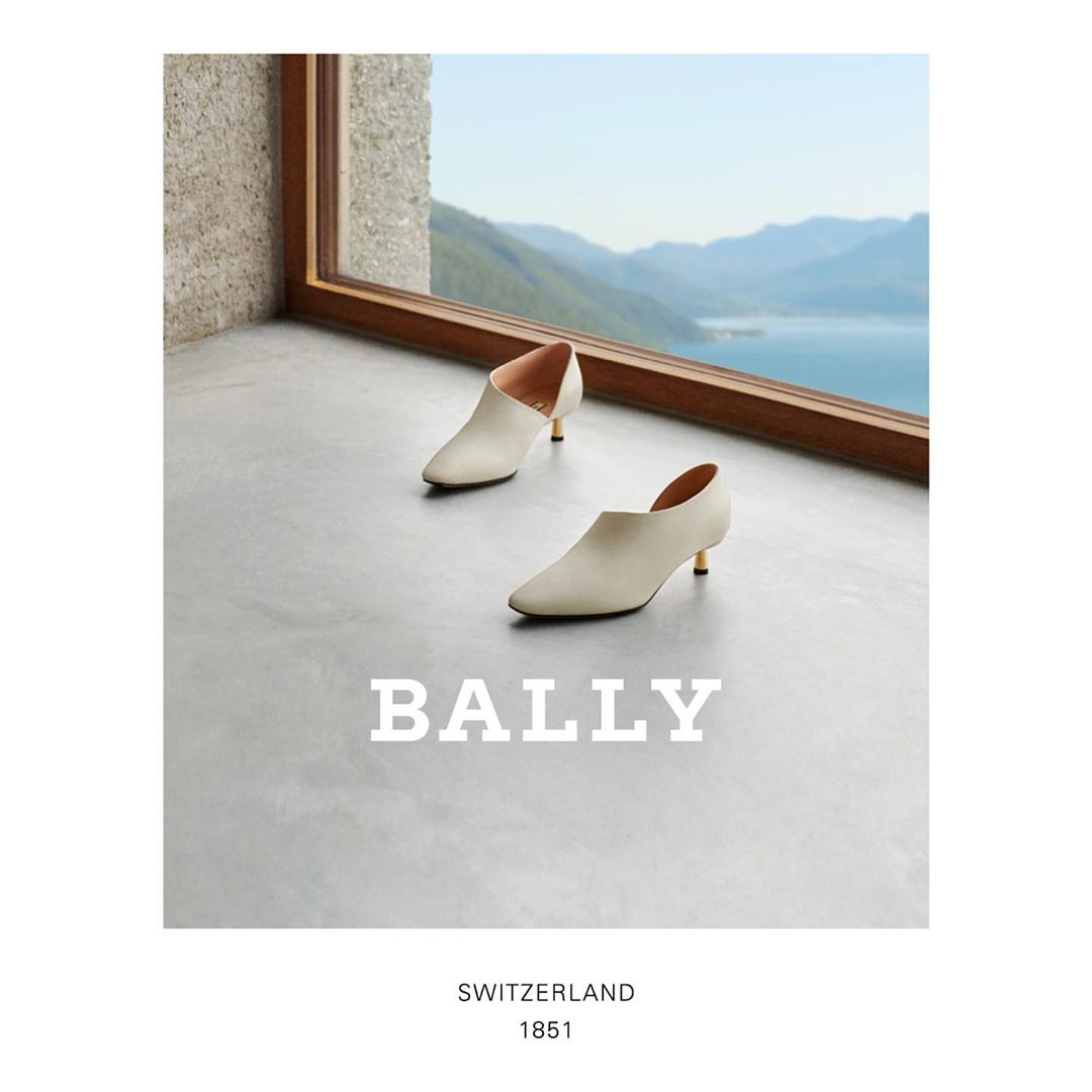 A pair of white Bally ankle boots with gold heels sit on a gray surface near a window with a view of a lake and mountains.
