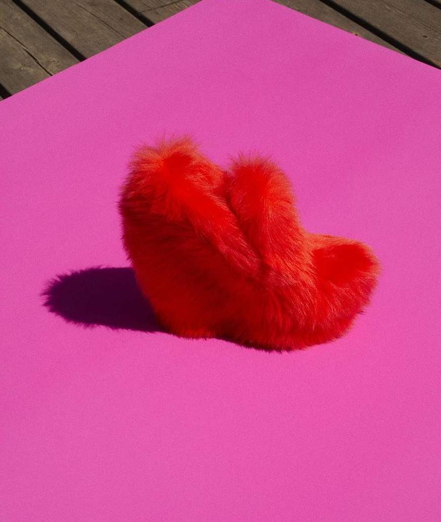 Fuzzy red slipper resting on a flat pink surface, studio shot with strong lighting and defined shadows, set against a light wood background.