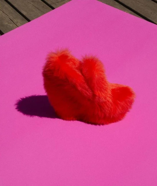 Fuzzy red slipper resting on a flat pink surface, studio shot with strong lighting and defined shadows, set against a light wood background.