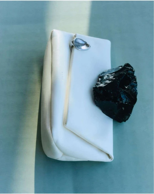 Still life shot of a white rectangular clutch with a silver fastener and a black mineral stone, presented on a blue-green surface.