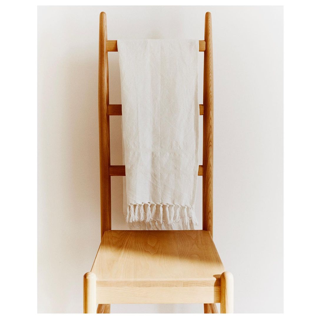 Still life of a wooden chair with a white linen towel draped over the back, indoors with neutral tones and soft, diffused lighting.