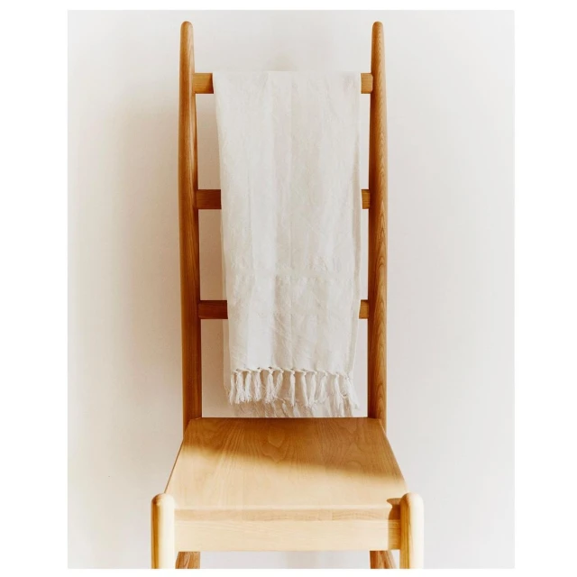Still life of a wooden chair with a white linen towel draped over the back, indoors with neutral tones and soft, diffused lighting.