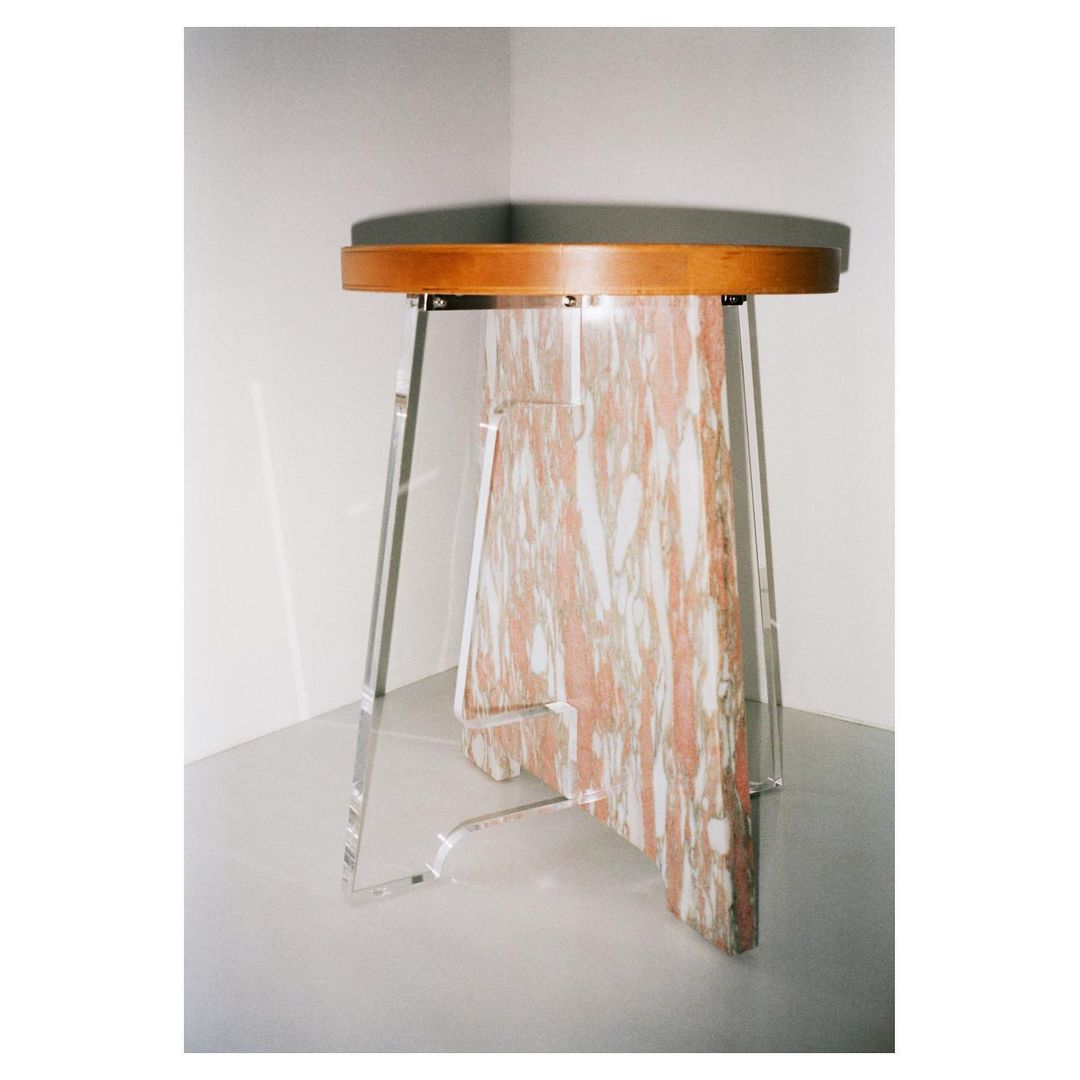 Modern accent table with wood top, marble center column, and clear acrylic legs sits against a white corner wall in an interior setting.