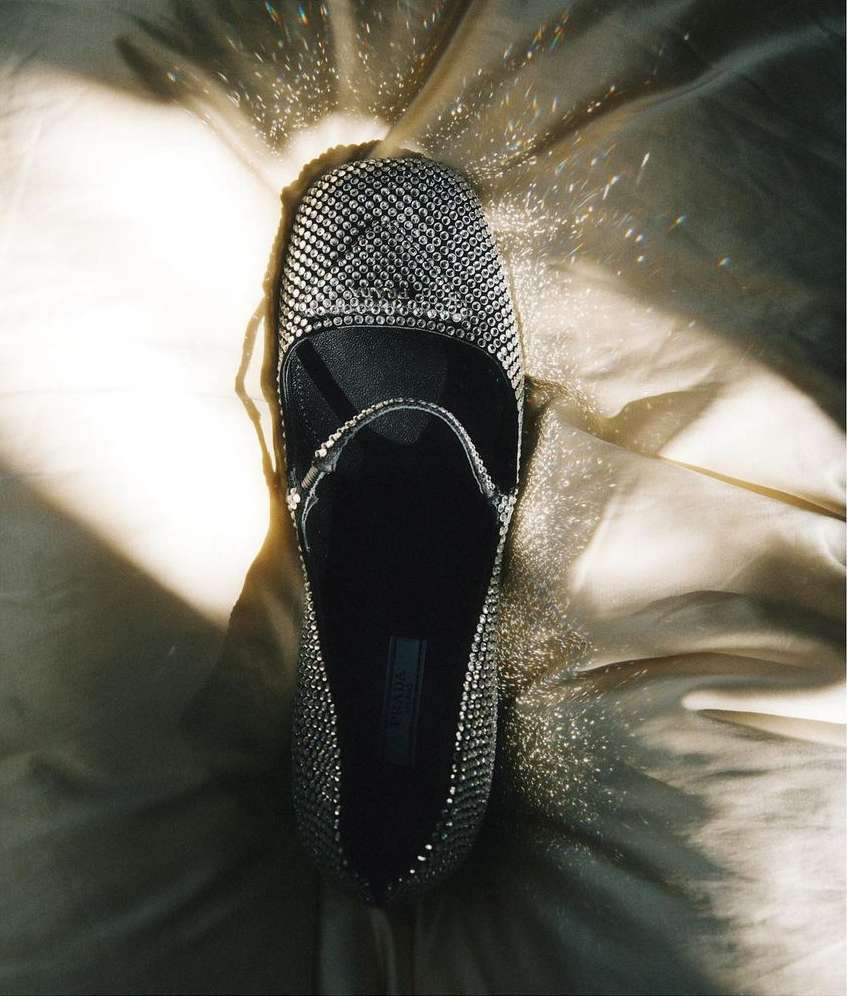 Close-up studio shot of a black, studded, Mary Jane style shoe on a shiny, light-gold satin-like fabric, lit from above creating contrast.