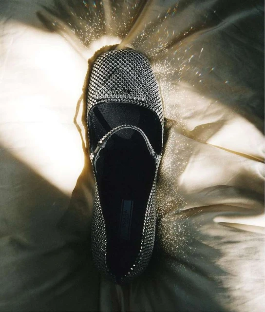 Close-up studio shot of a black, studded, Mary Jane style shoe on a shiny, light-gold satin-like fabric, lit from above creating contrast.