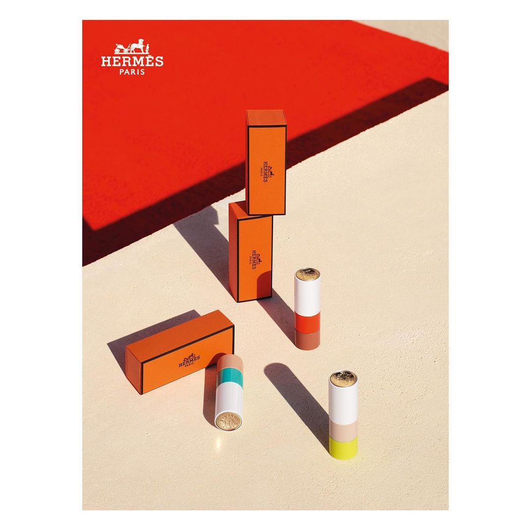 Hermes lipstick product photography featuring orange boxes, color-blocked lipstick tubes on a cream surface and a red background.