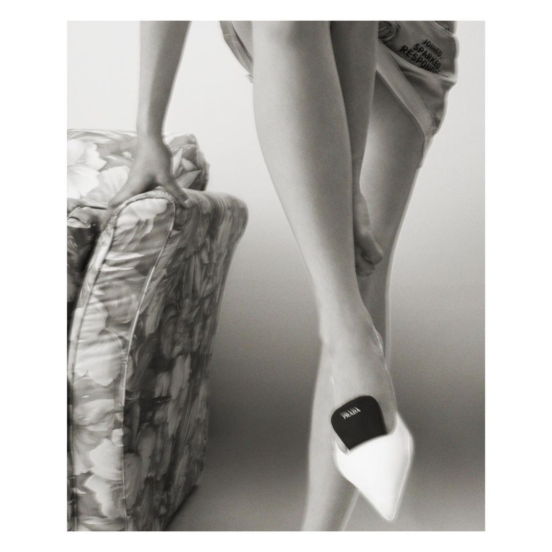Monochromatic fashion shot of a person wearing a pointed white high heel shoe and holding onto a patterned chair for support.