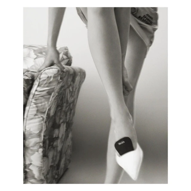 Monochromatic fashion shot of a person wearing a pointed white high heel shoe and holding onto a patterned chair for support.