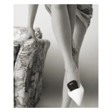 Monochromatic fashion shot of a person wearing a pointed white high heel shoe and holding onto a patterned chair for support.