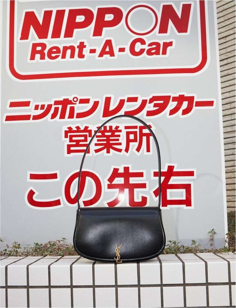 Elegant black YSL purse rests on a tiled wall, set against a sign with Japanese text and a logo. The bag is sleek and modern.