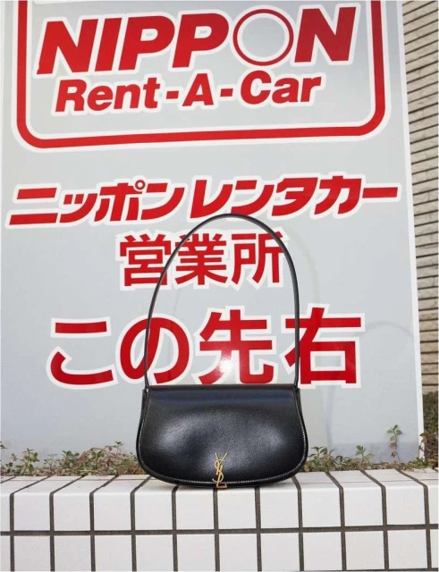 Elegant black YSL purse rests on a tiled wall, set against a sign with Japanese text and a logo. The bag is sleek and modern.