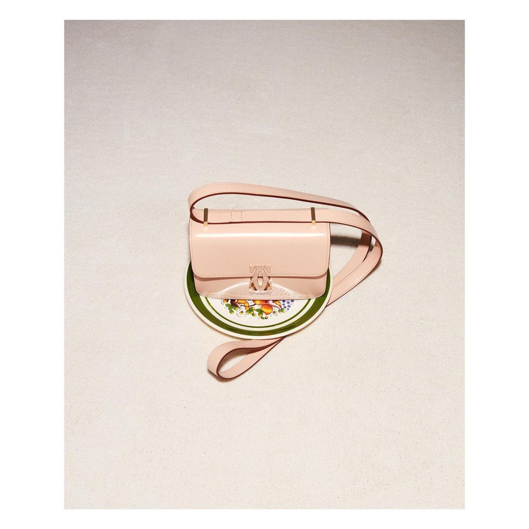 Glossy pale pink leather shoulder bag resting on a small decorative ceramic plate against a neutral textured background.