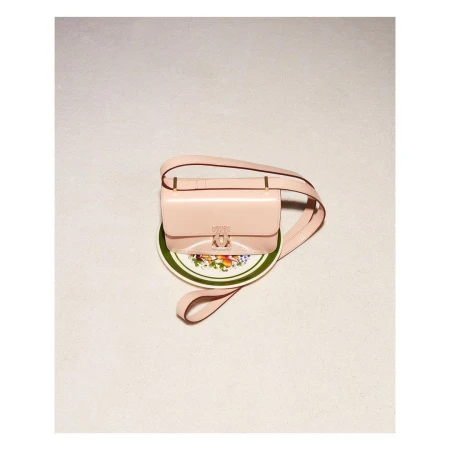 Glossy pale pink leather shoulder bag resting on a small decorative ceramic plate against a neutral textured background.
