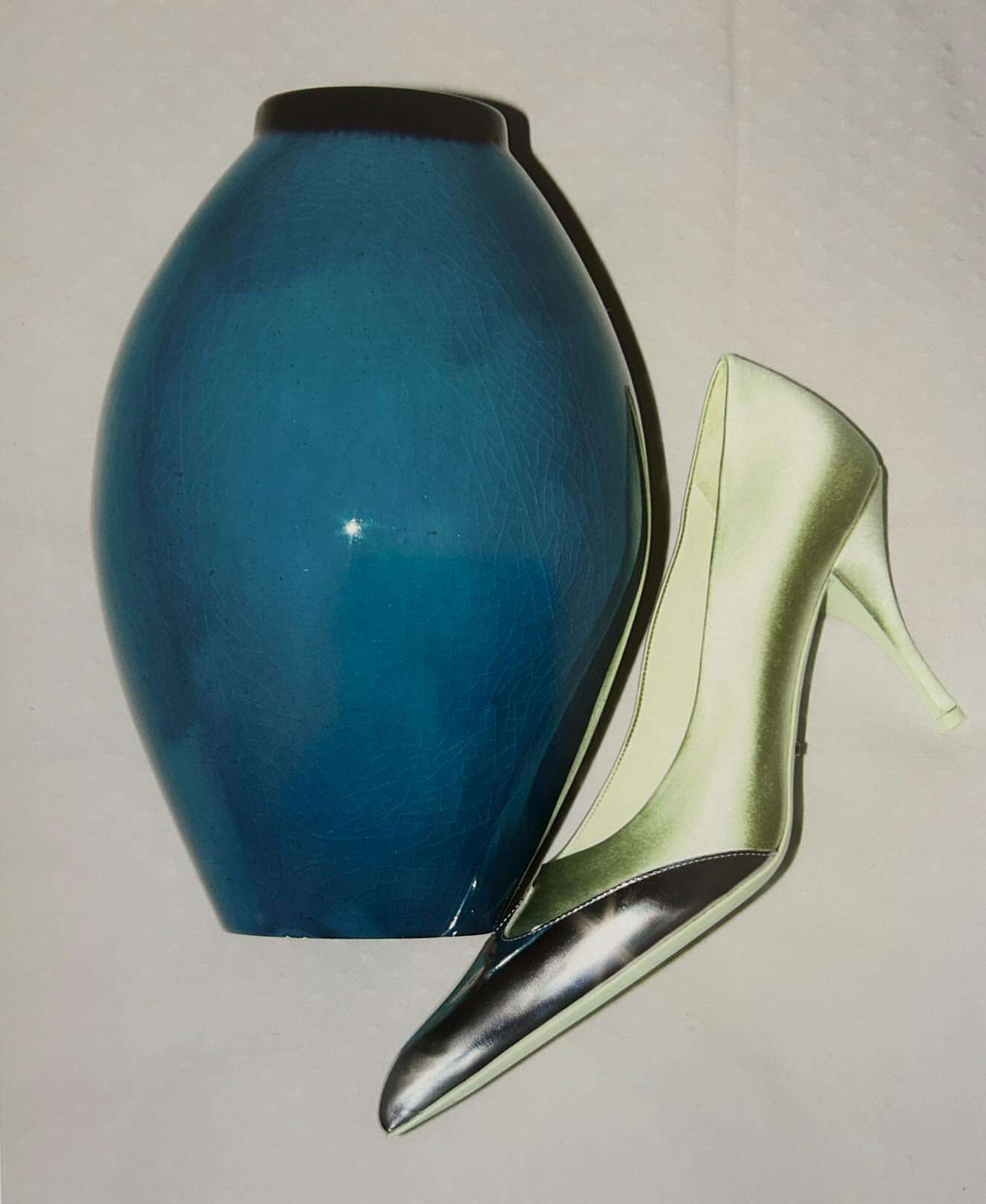 Turquoise vase with black rim and a single pale yellow high-heel shoe on a white background, studio lighting with soft shadows.