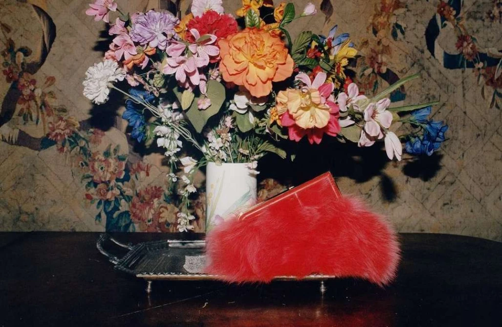 Still life with a colorful bouquet of flowers in a vase and a red furry clutch on a silver tray against a floral wallpaper backdrop.