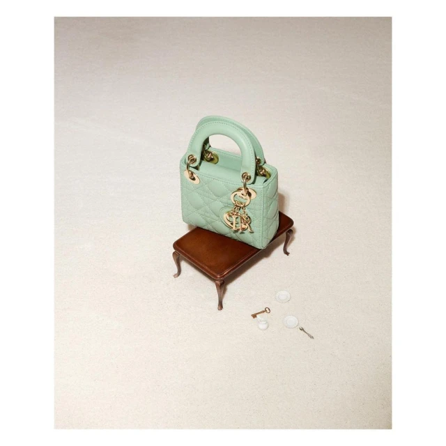 Mint green Lady Dior mini handbag on a tiny brown wooden table, studio shot. Gold logo charms add elegance to this luxury fashion accessory.
