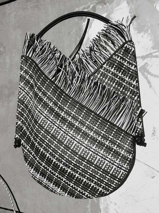 Woven tote bag with fringe detail and black handle, shot in black and white against a textured neutral background.