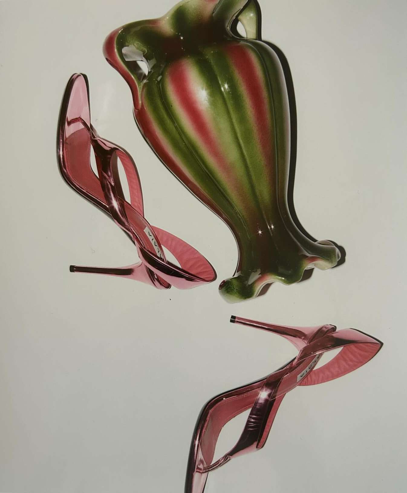 Still life composition featuring two high-heeled pink shoes and a green and pink striped vase against a white background.