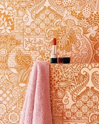 Close-up of a red lipstick standing beside its cap, paired with a textured pink towel against an orange and white patterned backdrop.