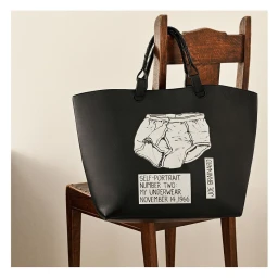 Black leather tote bag with illustrated underwear graphic hanging on the back of a wooden chair