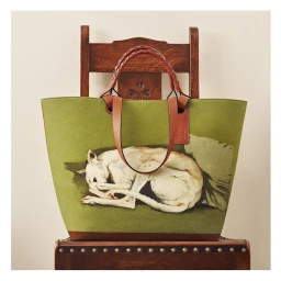 Green tote bag with leather handles and a sleeping white dog illustration, placed on a dark wooden chair.