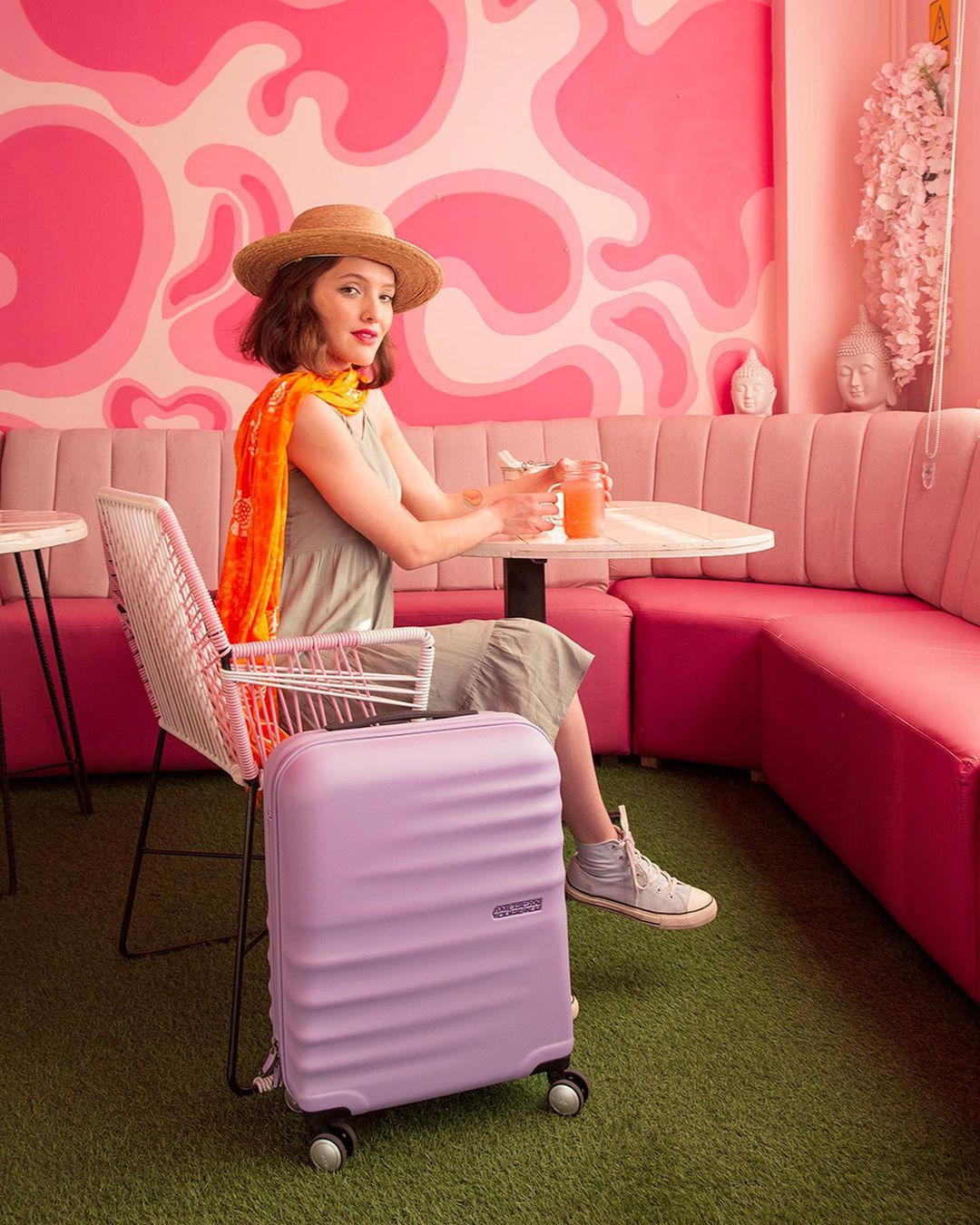 A young woman sits inside a pink cafe next to her purple suitcase, wearing a summer hat, scarf, and dress, ready for her travels.