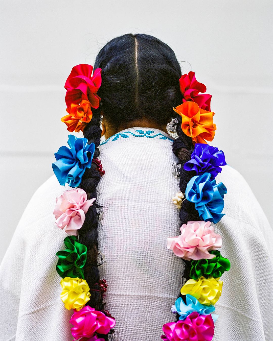 Back view of a person with dark hair styled in braids adorned with colorful flowers against a neutral background, fashion detail shot.