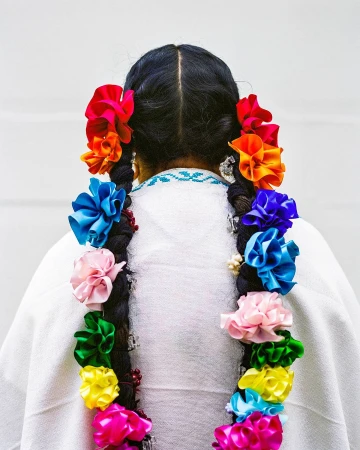 Back view of a person with dark hair styled in braids adorned with colorful flowers against a neutral background, fashion detail shot.