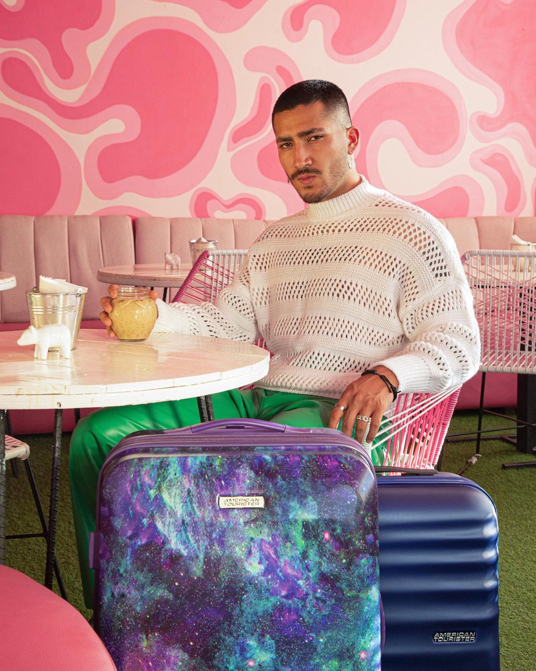 A man sits at a table in a cafe with pink walls and furnishings, two suitcases nearby. He is drinking a beverage.