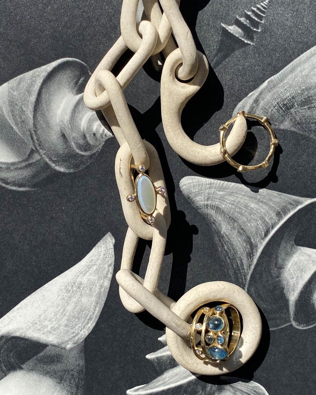 Jewelry still life featuring gold rings and a ceramic chain against a black and white seashell background in a studio setting.