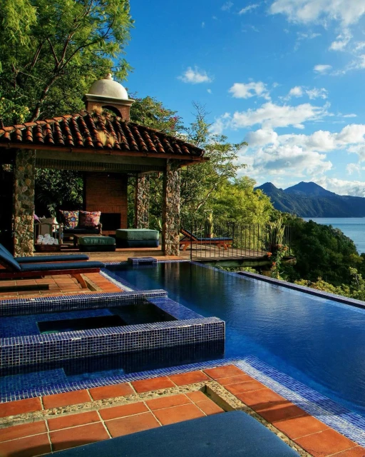 Luxury infinity pool overlooks lake and mountain backdrop from a stone gazebo and deck, under blue sky and cumulus clouds.