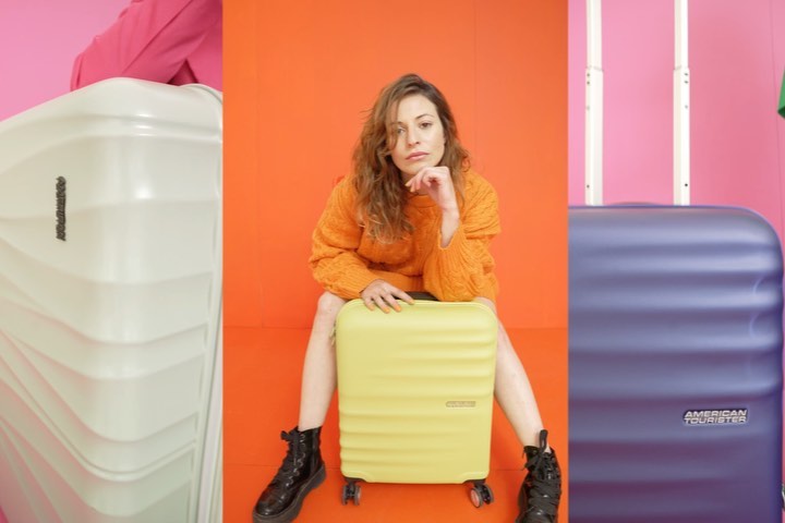 A young woman sits on a yellow suitcase in front of an orange background, flanked by a white suitcase on the left and a blue one on the right.