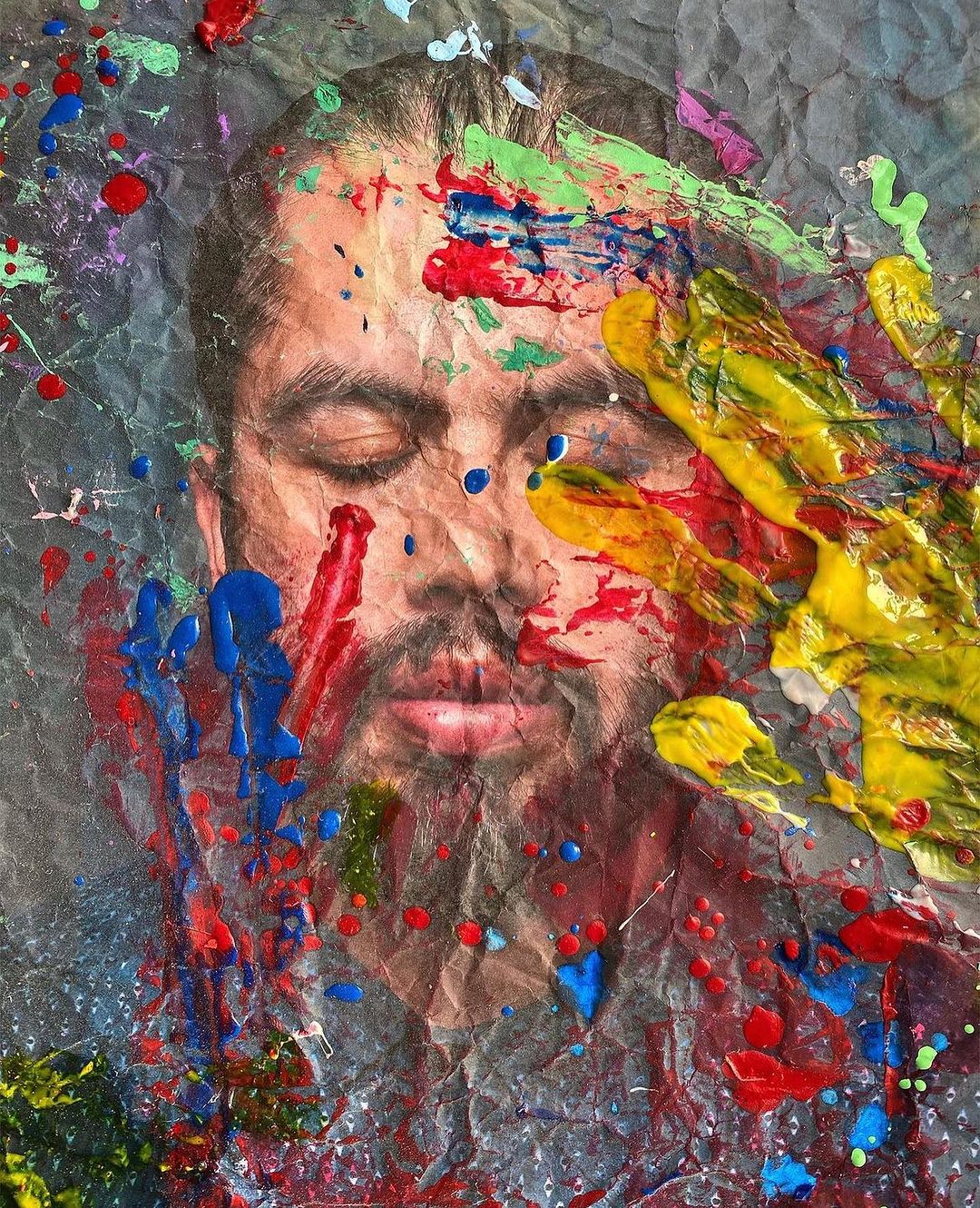 Painted portrait of an adult with eyes closed covered with bright paint splatters in red, green, yellow, and blue tones on gray paper.
