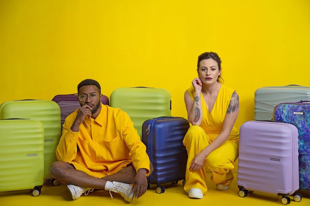 Two people in yellow outfits pose surrounded by colorful suitcases on a yellow backdrop, creating a vibrant and coordinated travel-themed image.