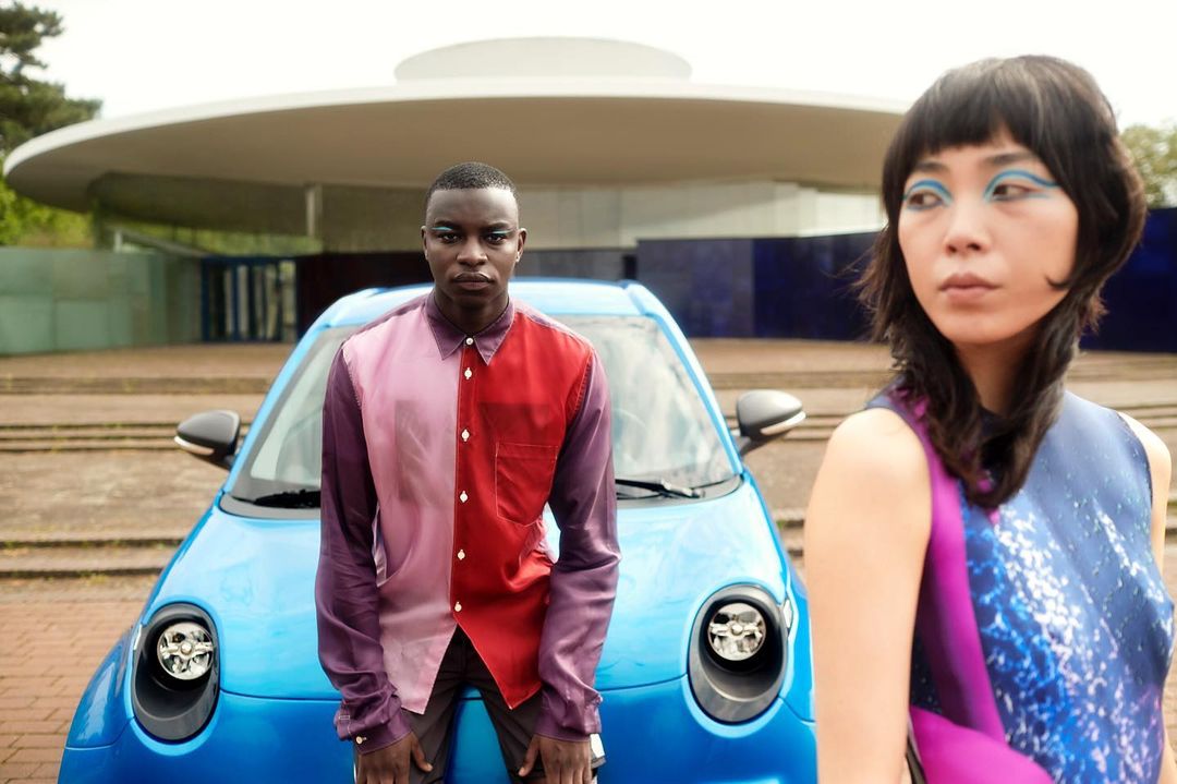 Two fashion models posing outdoors next to a blue car and modern architecture, showcasing colorful clothing and bold eye makeup.