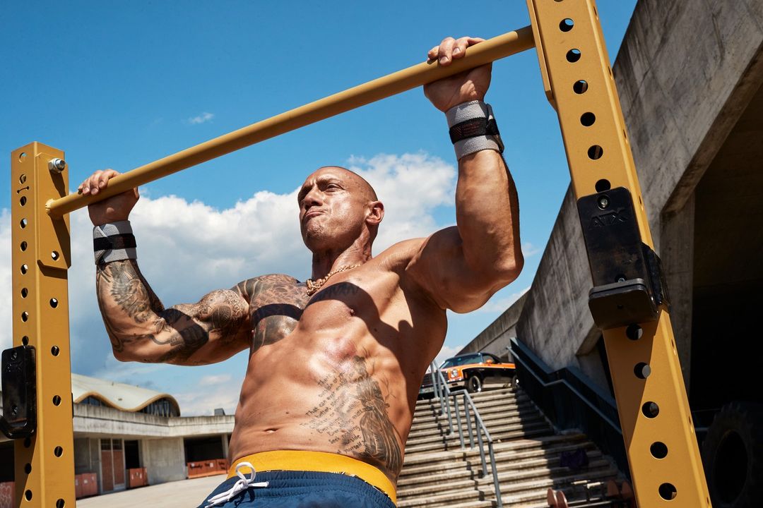 Muscular man with tattoos doing pull-ups outdoors on sunny day, showcasing strength and fitness in an urban setting.