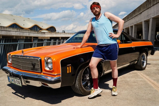 Man in retro athletic wear poses with a black and orange classic muscle car on concrete with modern architecture in background.
